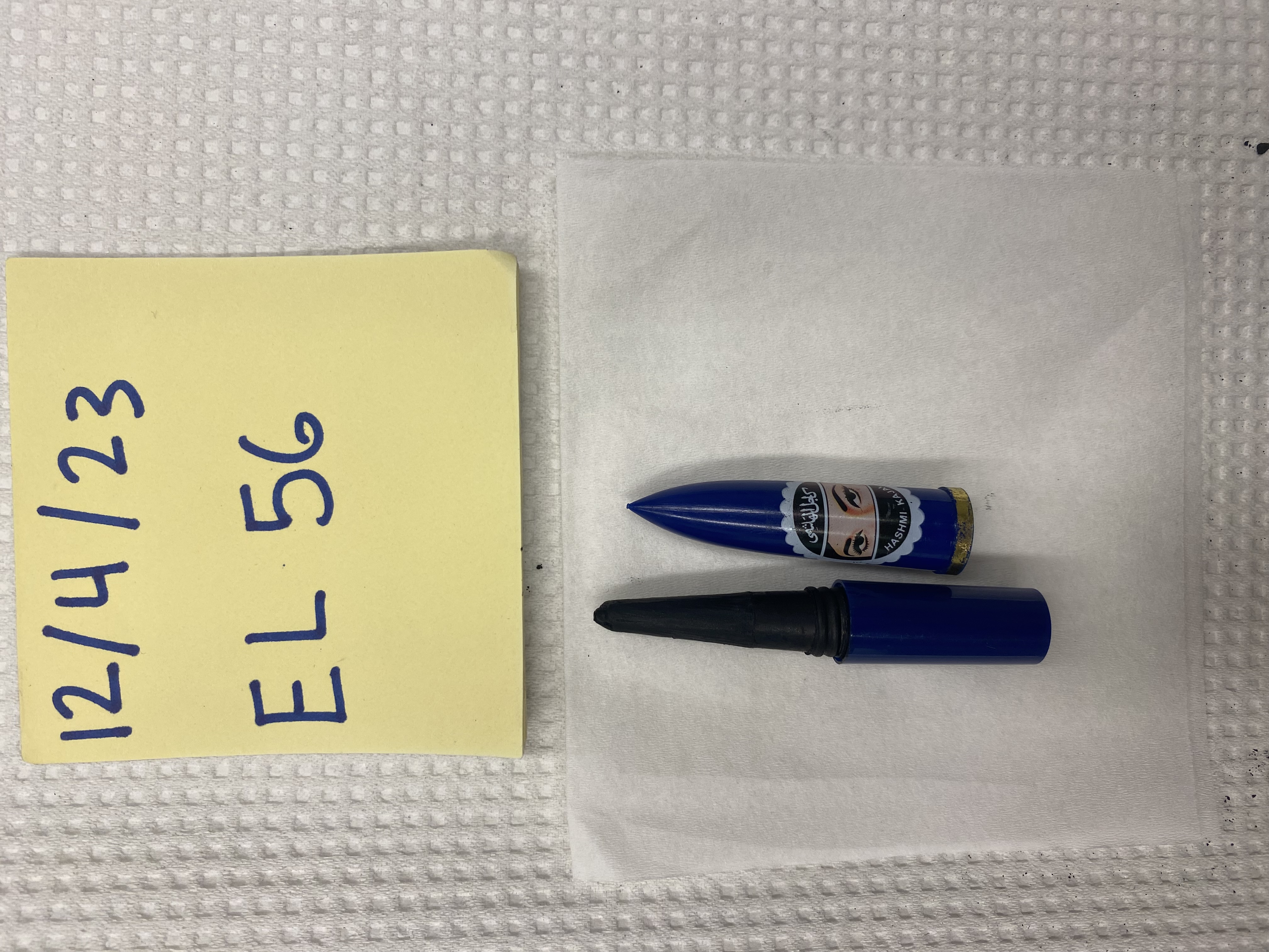 "A bottle of imported eyeliner with a wand applicator. The cap is sitting next to the wand and has a picture of a woman with a headscarf and Arabic text. Near the eyeliner is a post-it note reading 12/4/23 EL 56.