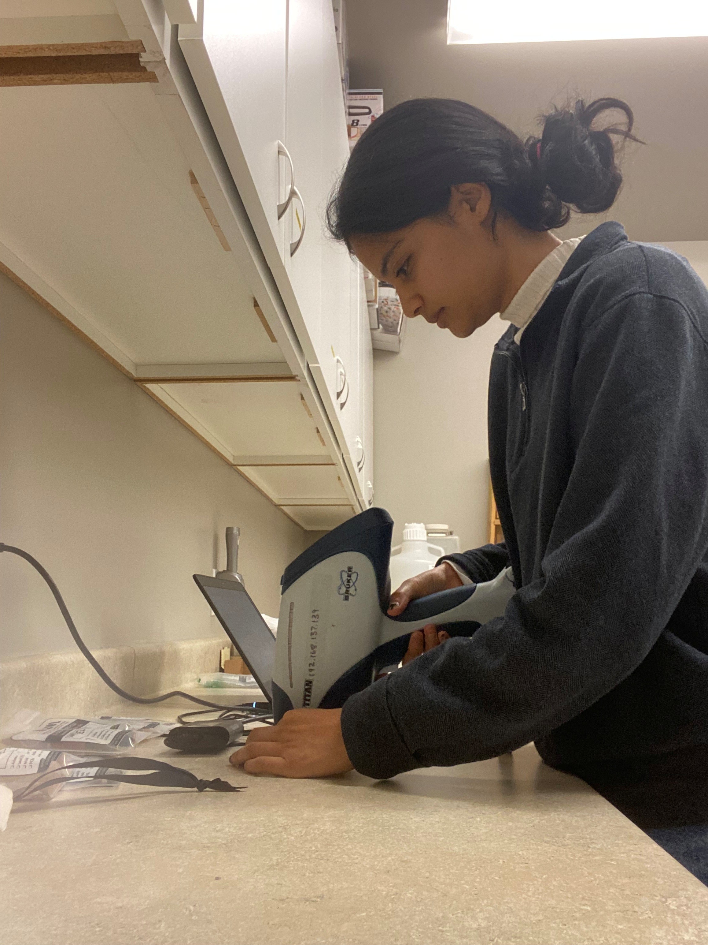 Mokashi stands at a lab bench holding an X-ray fluorescence analyzer over an eyeliner sample.