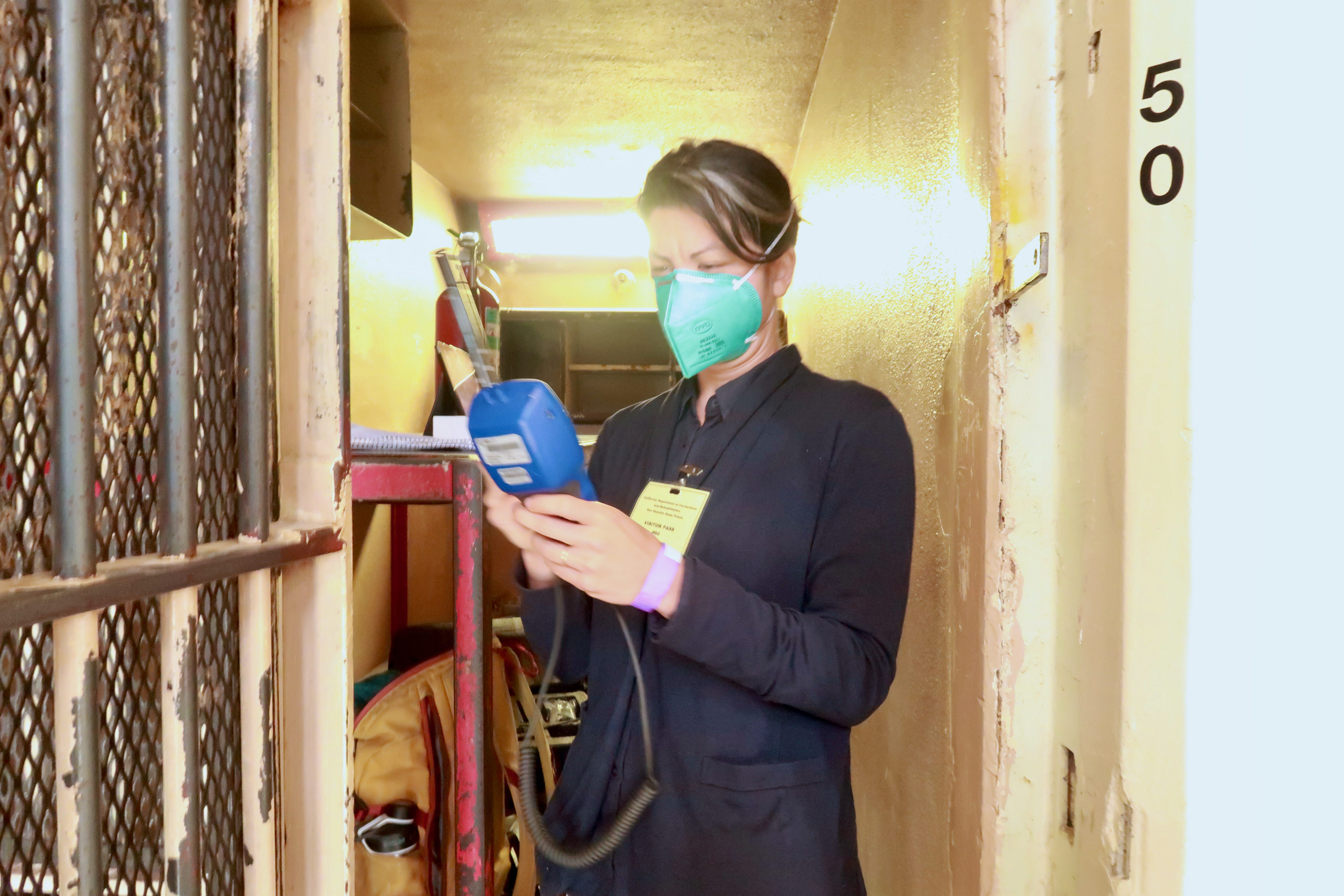 Sklar stands in a prison cell with a respiratory mask holding an air monitoring device.