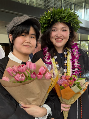 Two graduates smile holding bouquets of flowers.