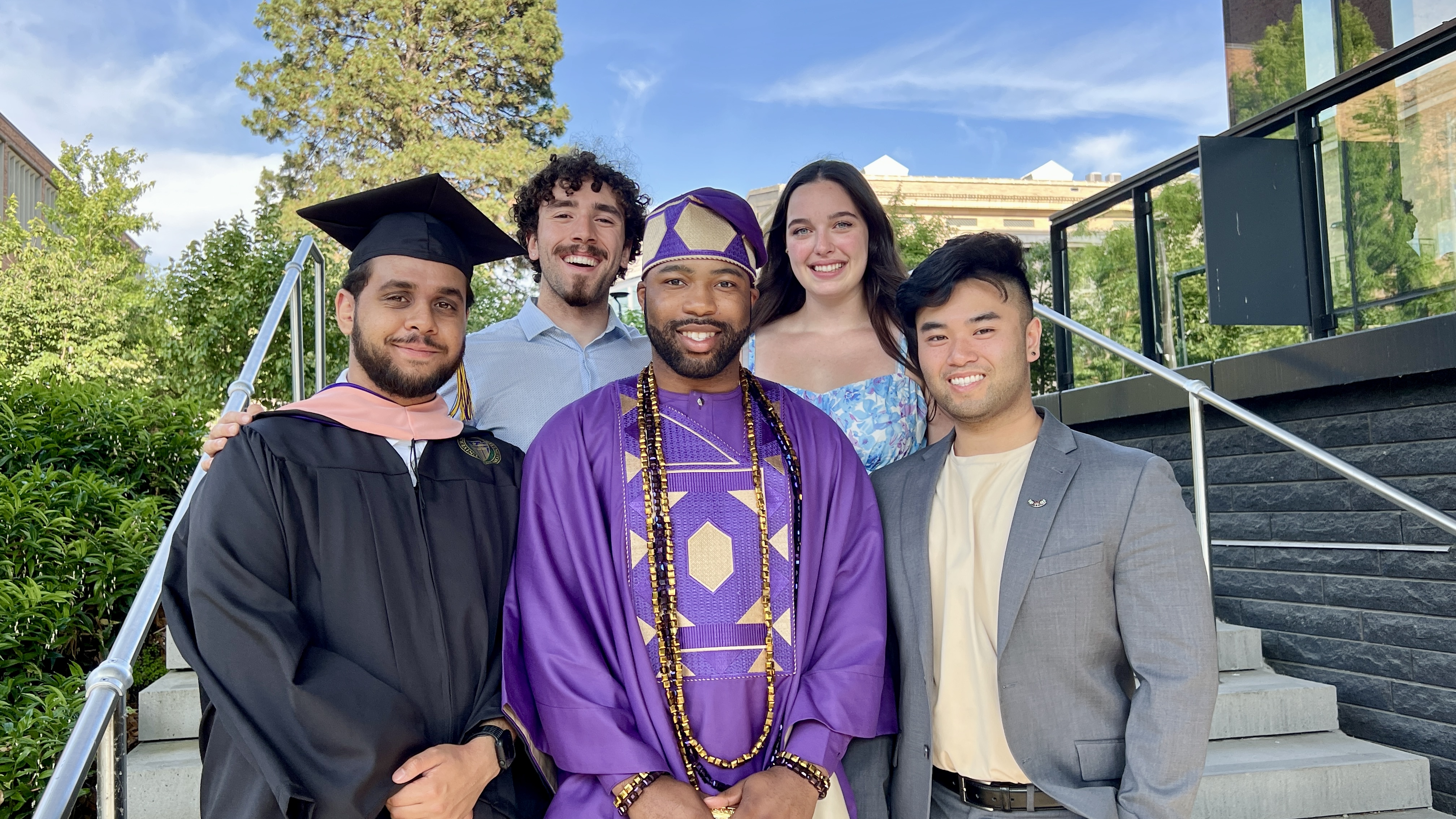 Five students stand together on steps on the UW campus, with one on the left wearing a cap and gown.