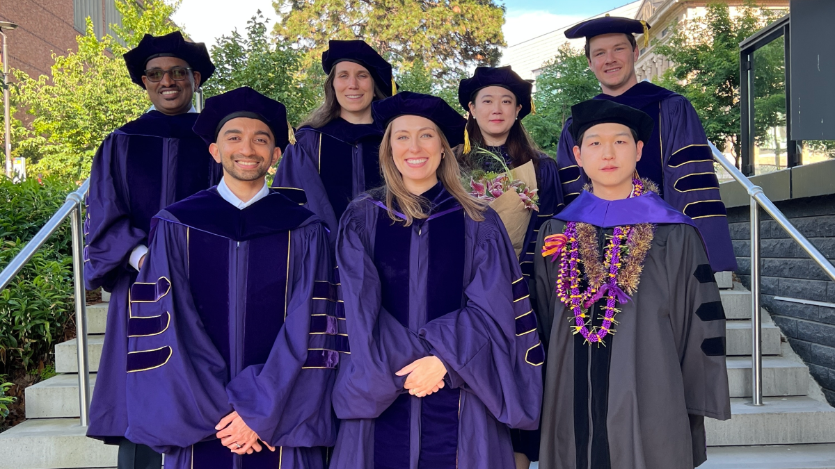 Seven PhD students stand together on the steps of the UW campus wearing regalia.