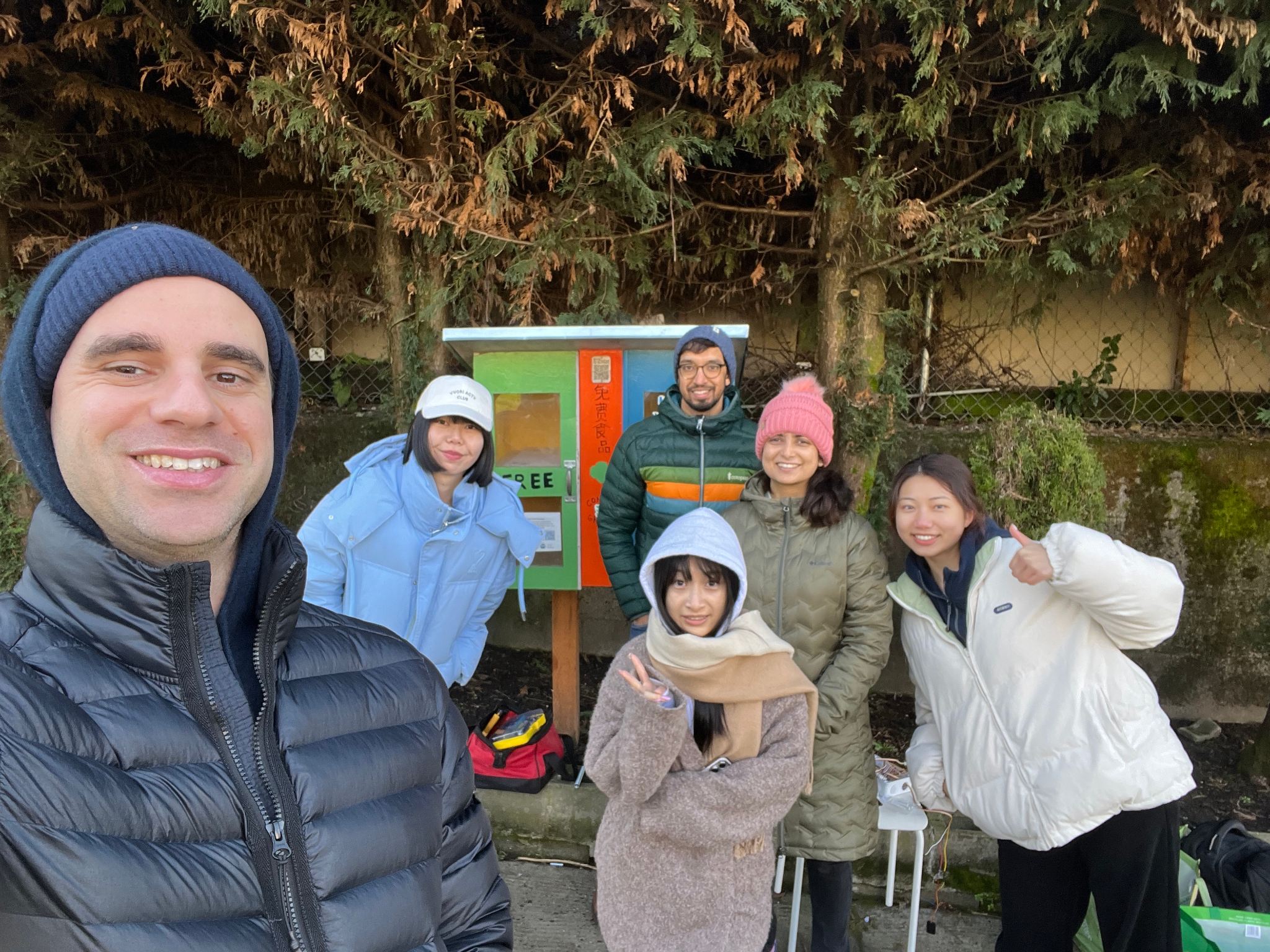 Six people stand in front of a micropantry with free food.