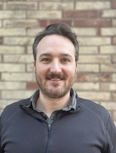 Image of a smiling man with a grey sweatshirt standing in front of brick wall