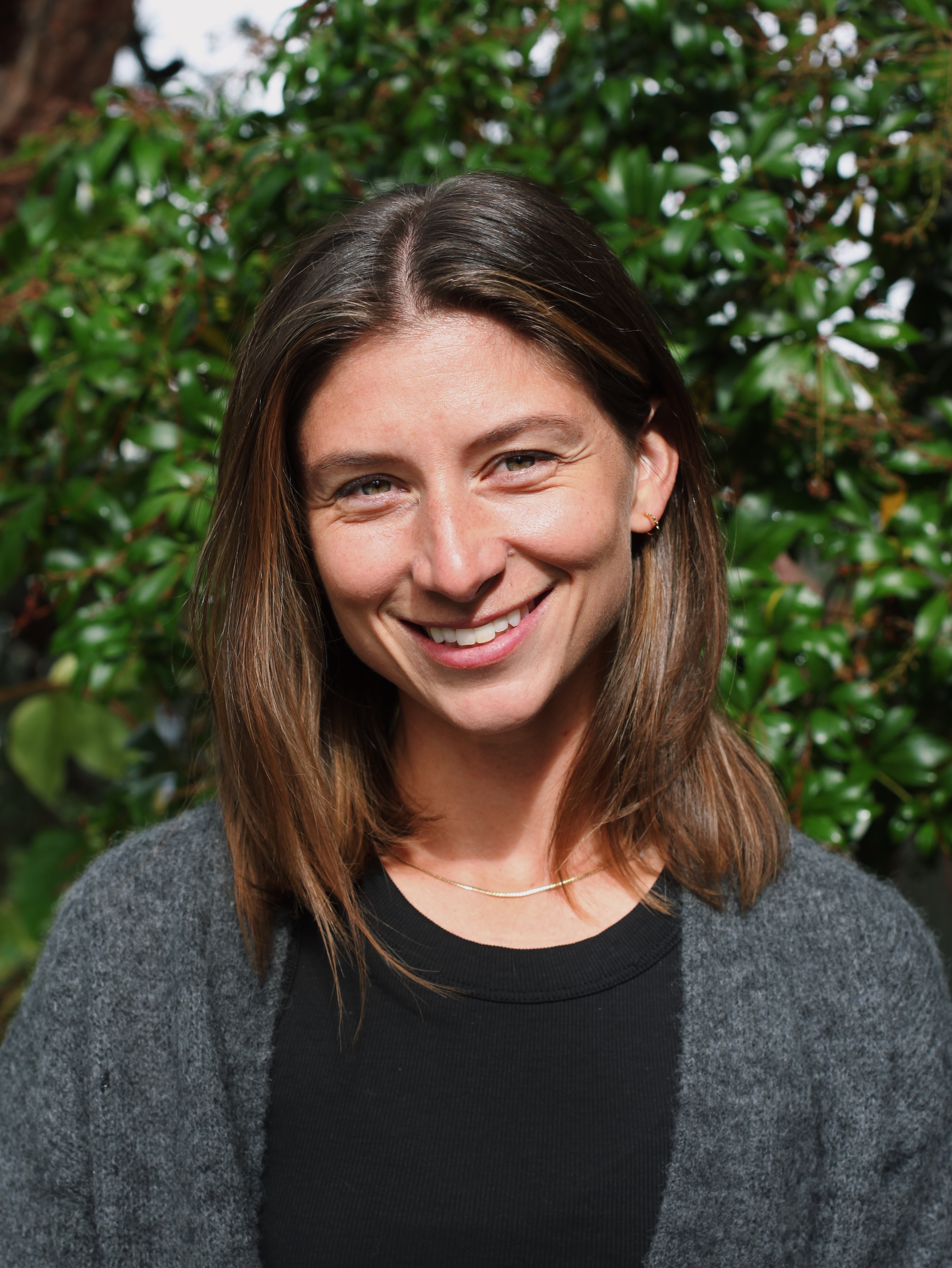 A smiling woman with brown hair standing in front of green leaves. 
