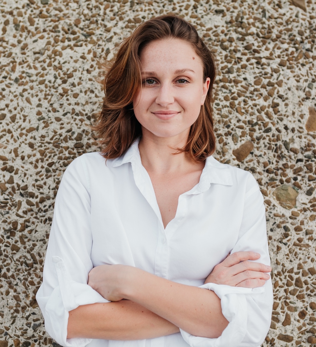 A smiling woman with brown hair is wearing a white shirt and standing in front of a tan stone wall.