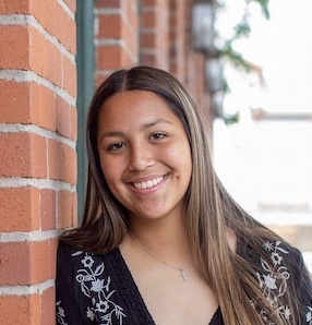 A young woman with brown hair and a black shirt with white embroidery is smiling and leaning against a brick wall.