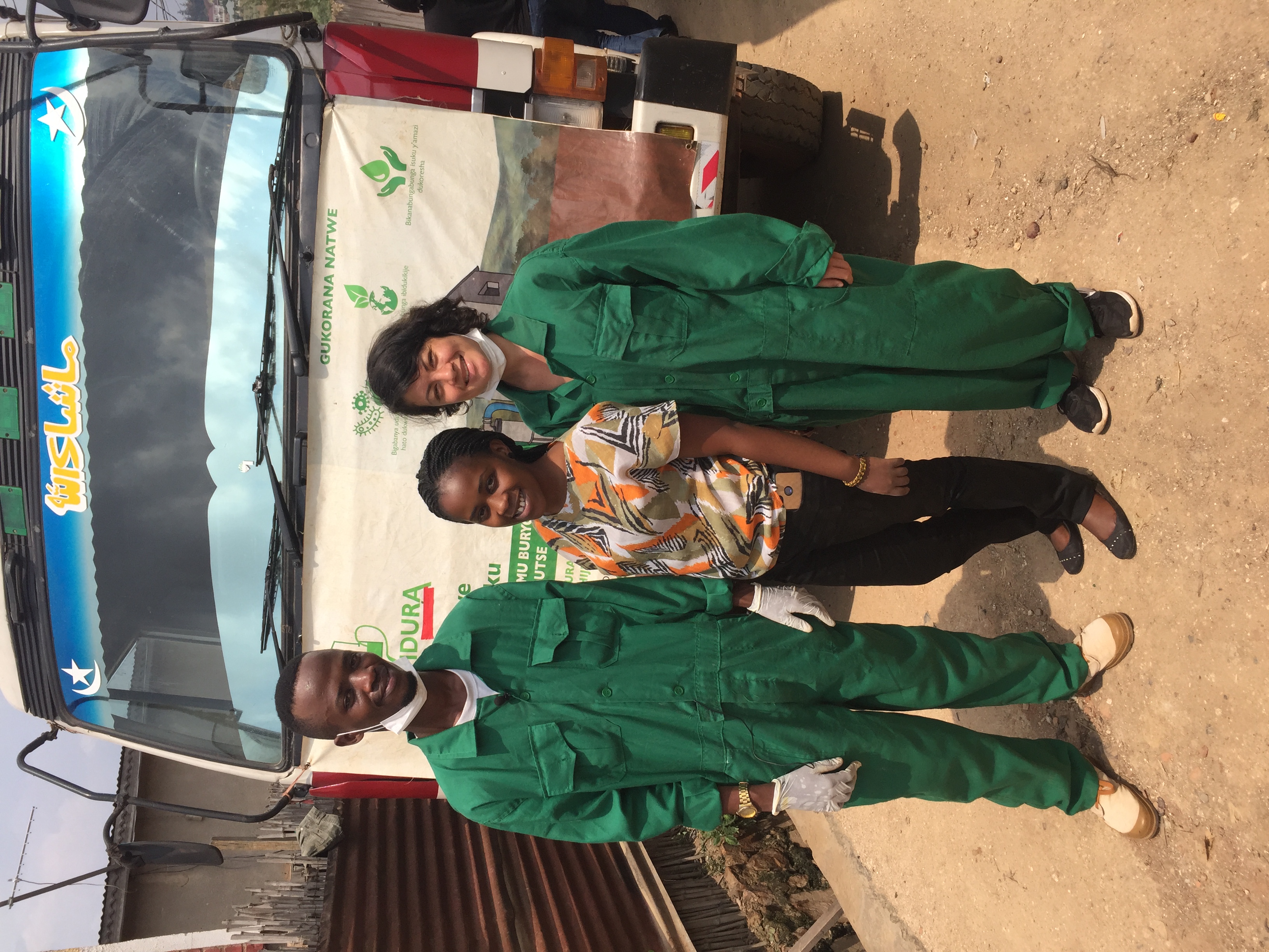 Sklar stands with two sanitation workers in front of a truck on the street in Kigali. Sklar and one worker are wearing green jumpsuits, the other worker is wearing a brightly colored blouse and black pants.