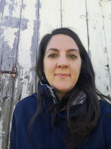 Woman with long black hair wearing dark blue jacket against a background of wood painted white