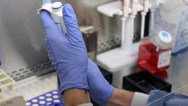 Two hands with blue medical gloves hold a medical device. Lab equipment out of focus is in the background.
