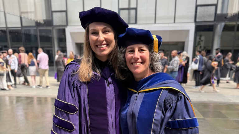 Rebecca Kann and Karen Levy stand together smiling wearing purple graduation regalia.