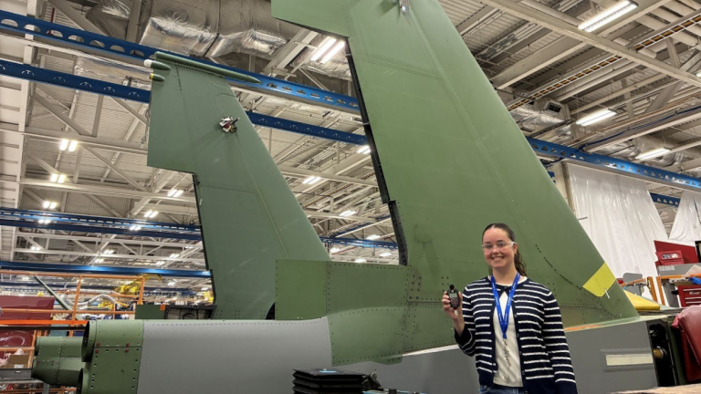 Lauren Costa stands in front of a military aircraft holding a noise monitor.