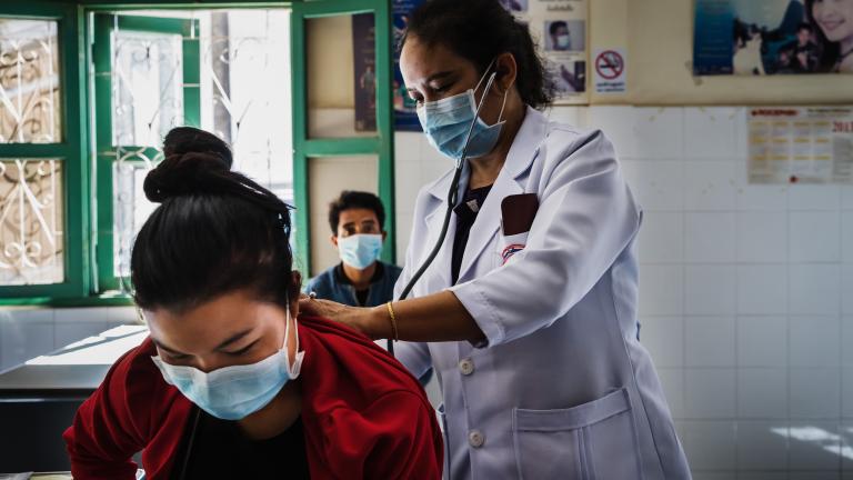 A healthcare worker stands behind a patient listening to the patient's lungs with a stethoscope while the patient leans over and looks down. Another person is sitting along a wall behind the two.
