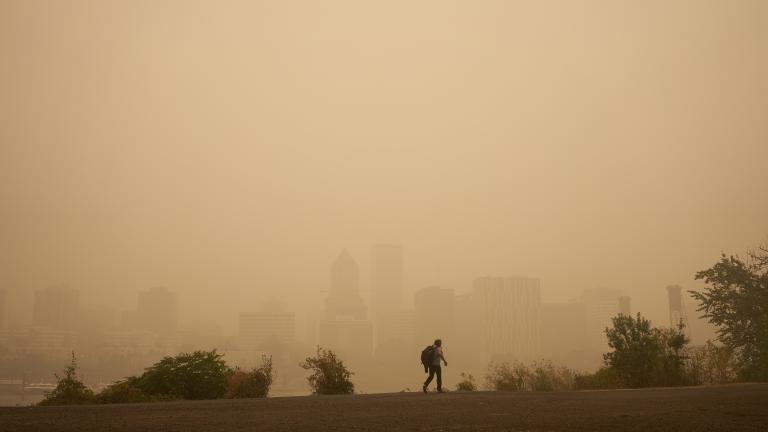 Smoky skies turn the Portland skyline hazy and orange-hued. A person with a backpack walks in the foreground of the image.