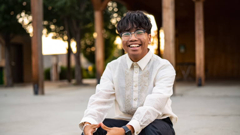 Elijah Morales smiles while sitting outside on steps in front of columns in the background.
