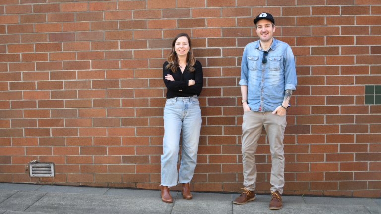 Kathleen Moloney and Evan Mix stand outside with their backs against a brick wall.