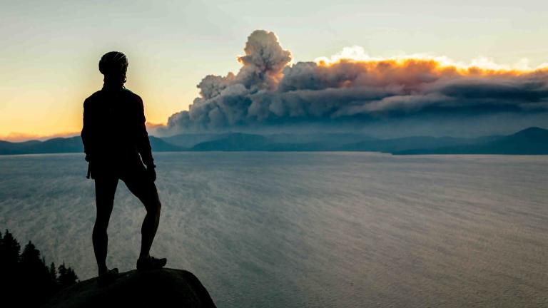 A woman in a bike helmet looks out over a lake toward wildfire smoke in the distance