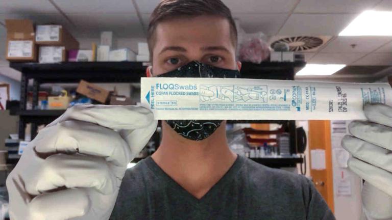 A man stands in a lab holding a swab in its paper packaging.
