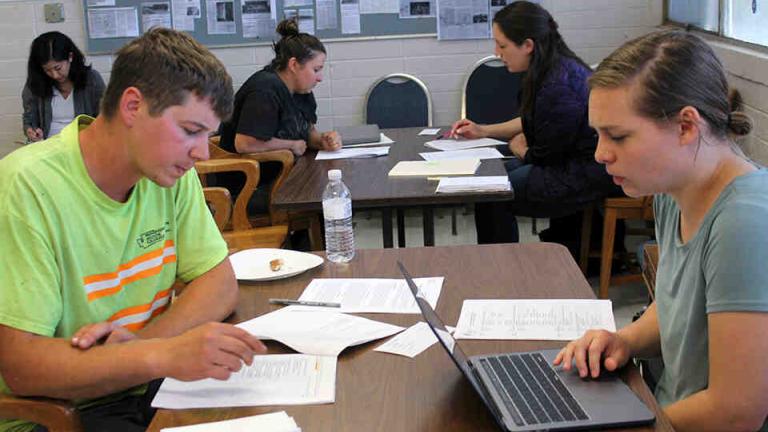 Students sit at tables interviewing construction workers.  In the foreground a female student with a laptop interviews a male construction worker with a packet of papers in front of him.