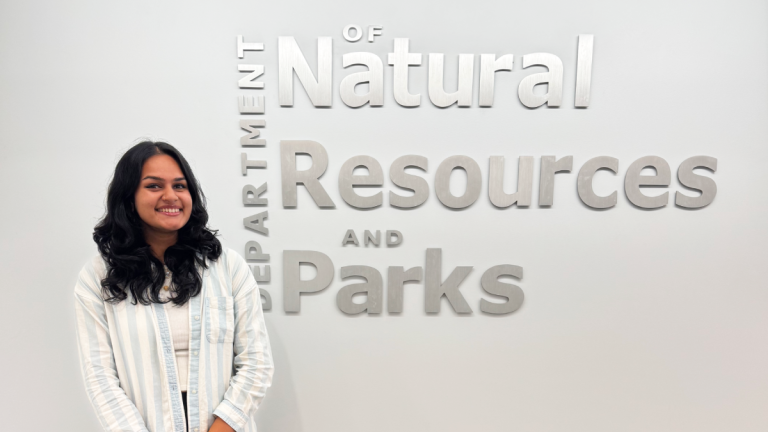 Tejasvini Vijay stands in front of a white wall with silver letters reading "Department of Natural Resources and Parks"