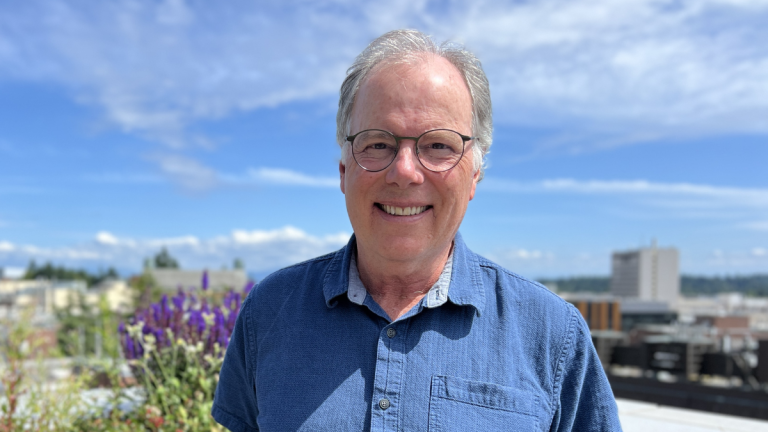 Jerry Cangelosi smiles while standing outside on a roof deck, with flowers and buildings in the background.