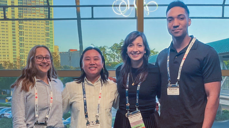 Four people (Hannah Dye, Jenielle Domaoal, Judit Marsillach and Elijah Scott) stand together smiling inside in front of a window, with a tall building outside in the background.