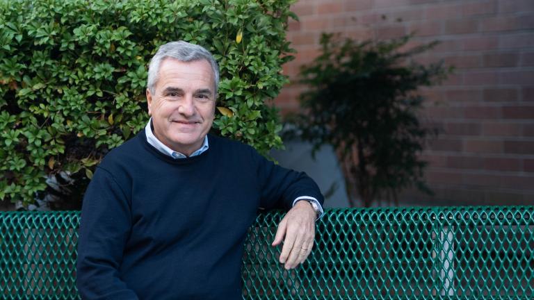 A man wearing a blue sweater sits on a green park bench against a backdrop of greenery and brick wall.