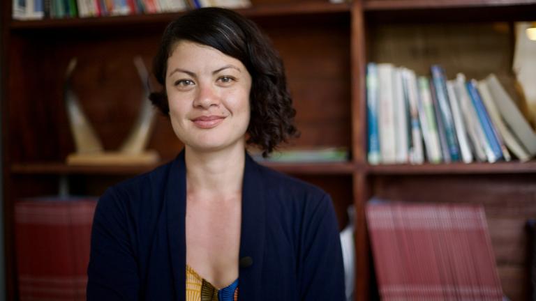 Rachel Sklar smiles while standing in a room with a bookshelf of books behind her.