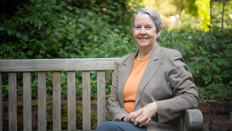 Lianne Sheppard sits on a wooden bench in front of green trees and shrubs. She wears a light orange shirt and light brown jacket with a pearl necklace and has her hands folded in her lap while smiling at the camera.