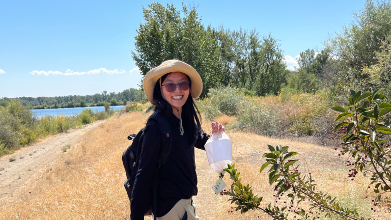 Sophia Li smiles wearing sunglasses and a hat and holding a mosquito trap outside with a dirt trail, trees and bushes, and a lake in the background.