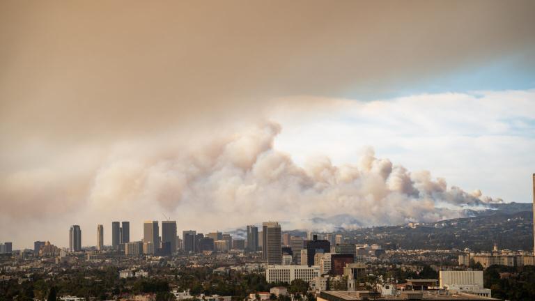 Skyscrapers of Los Angeles in the foreground with large plumes of smoke rising up high into the sky in the background.