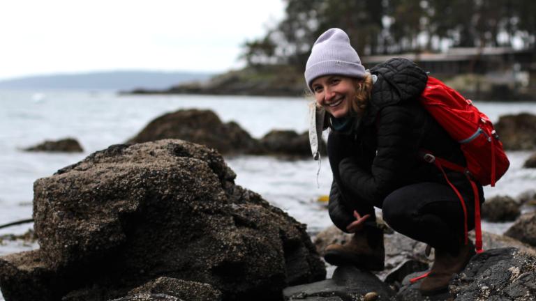 Person dressed in black wearing a purple hat and red backpack crouches on a rocky shoreline with trees in distance.