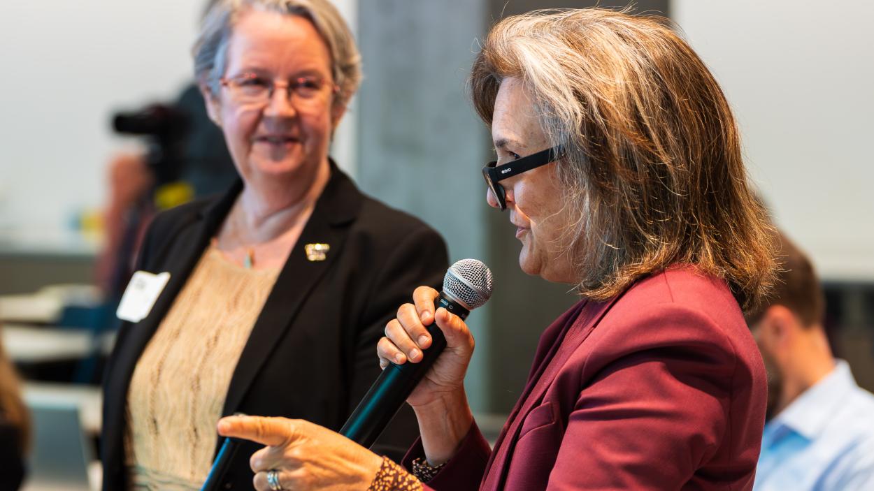 Tracey Woodruff speaks into a microphone while Lianne Sheppard looks on.