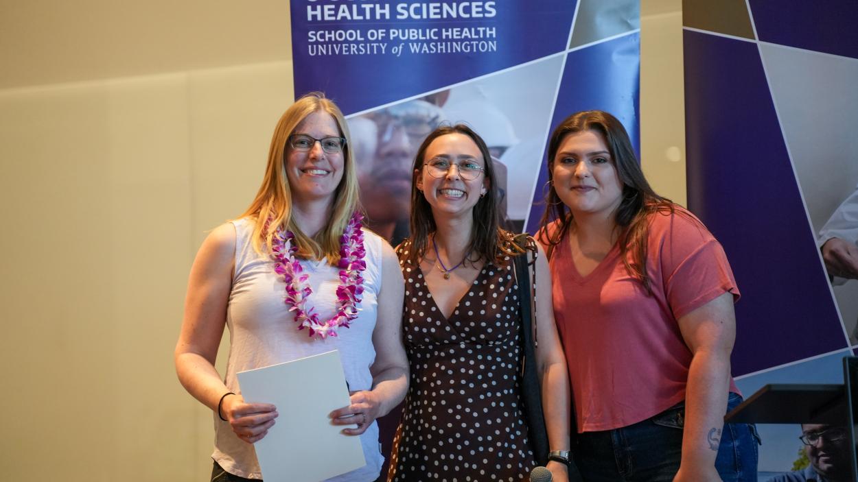 Emily Hovis, Mariah Rubin and Gracie Hively smile together on a stage with a banner reading Department of Environmental & Occupational Health Sciences behind them.