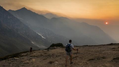 A smoky sunset with mountains and a valley in the background and a hiker facing away in the foreground.