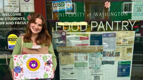 Angelina Durbin stands in front of the window of the UW Food Pantry. She is holding a cardboard box reading "No Husky Goes Hungry" with an image of a husky dog.