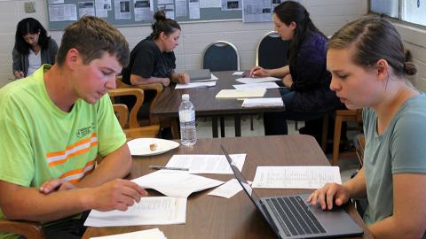 Students sit at tables interviewing construction workers. In the foreground a female student with a laptop interviews a male construction worker with a packet of papers in front of him.