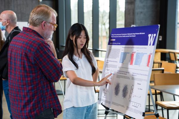 A person points to a research poster on an easel while another person looks on.